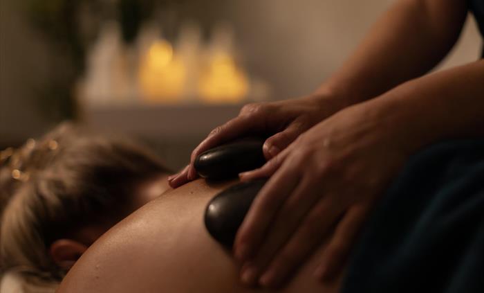 Image shows a guest receiving a relaxing hot stone spa treatment in a dimmed room, lit with candles.